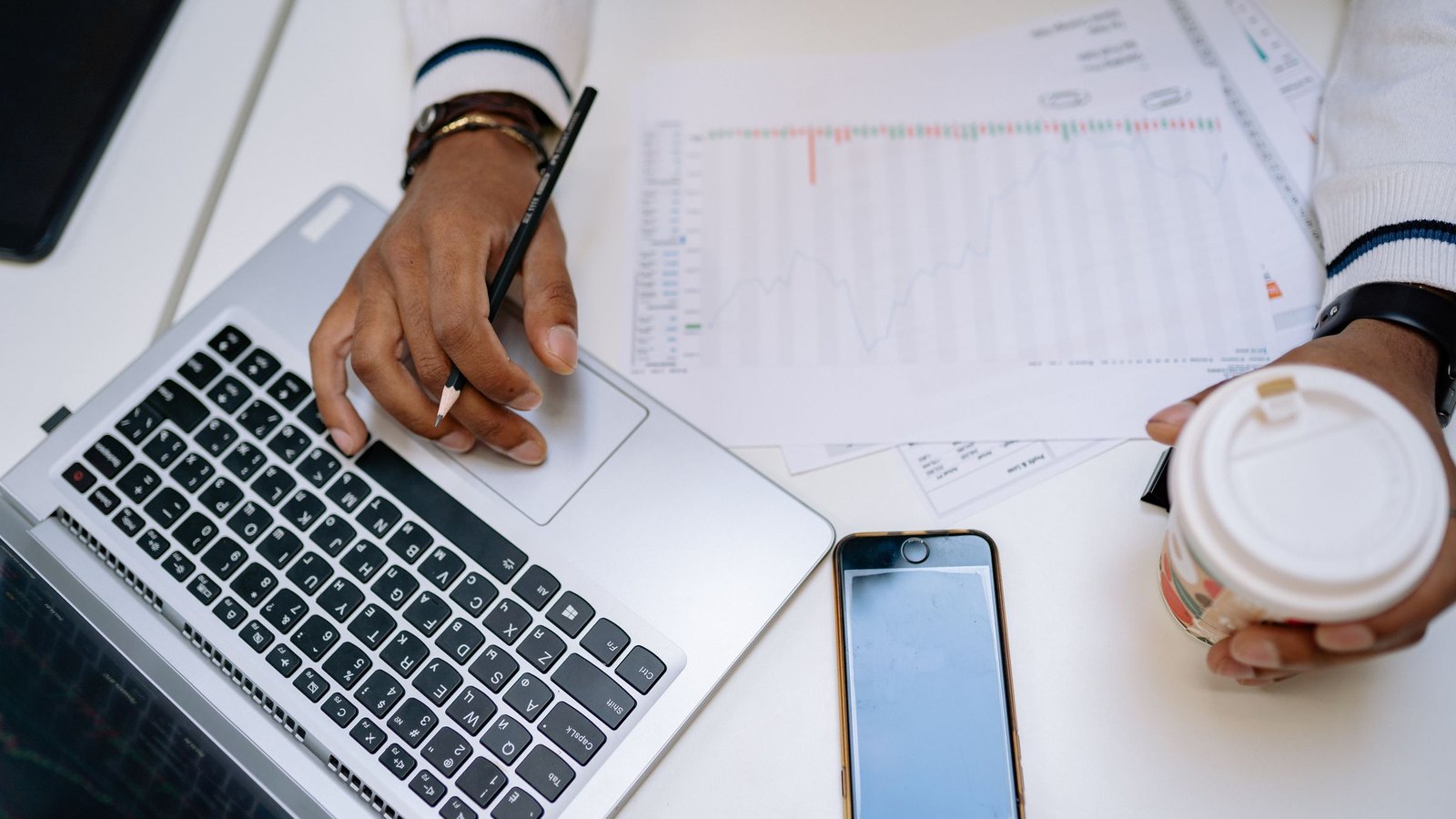 Finance lead comparing a printed cost sheet with figures on a laptop, phone and coffee at hand