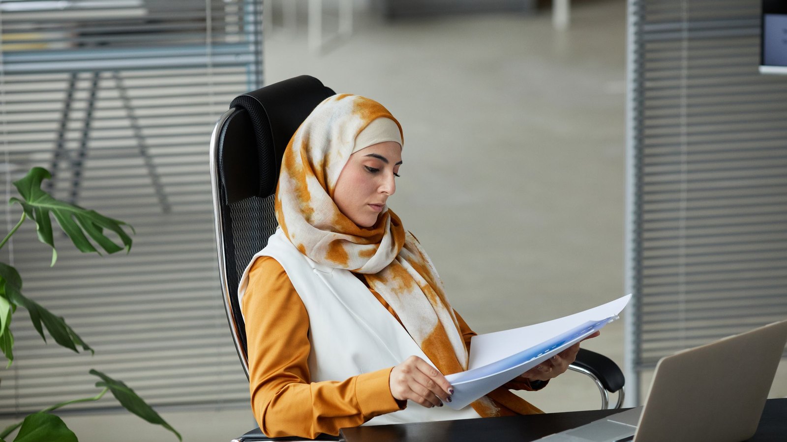 Finance officer in a hijab reviewing a printed invoice against her laptop to flag missing buyer details