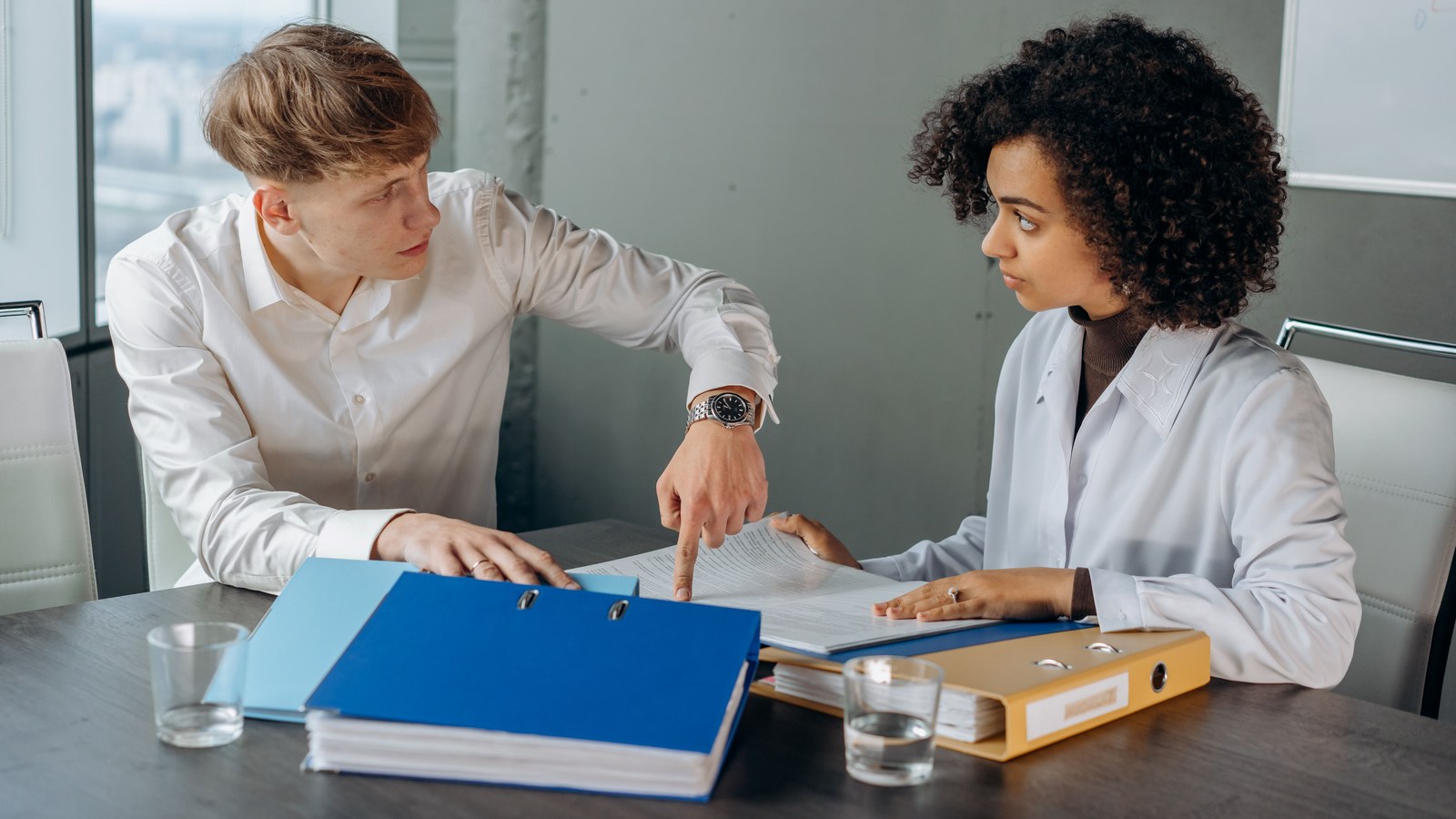 Two colleagues going through separate blue and yellow binders of company records at an office meeting table
