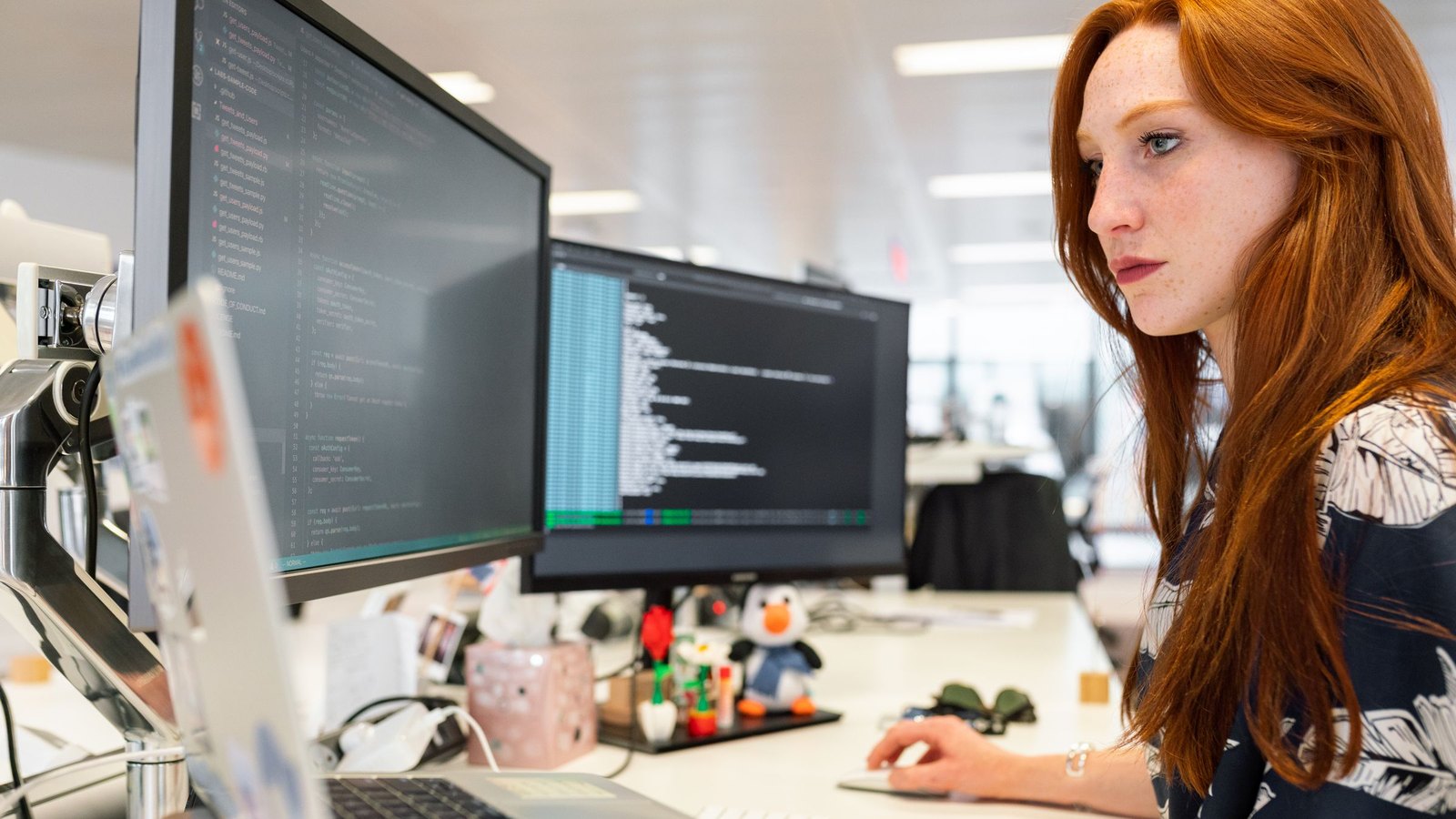 Software engineer at a desk of monitors showing terminal output and code, working on a laptop