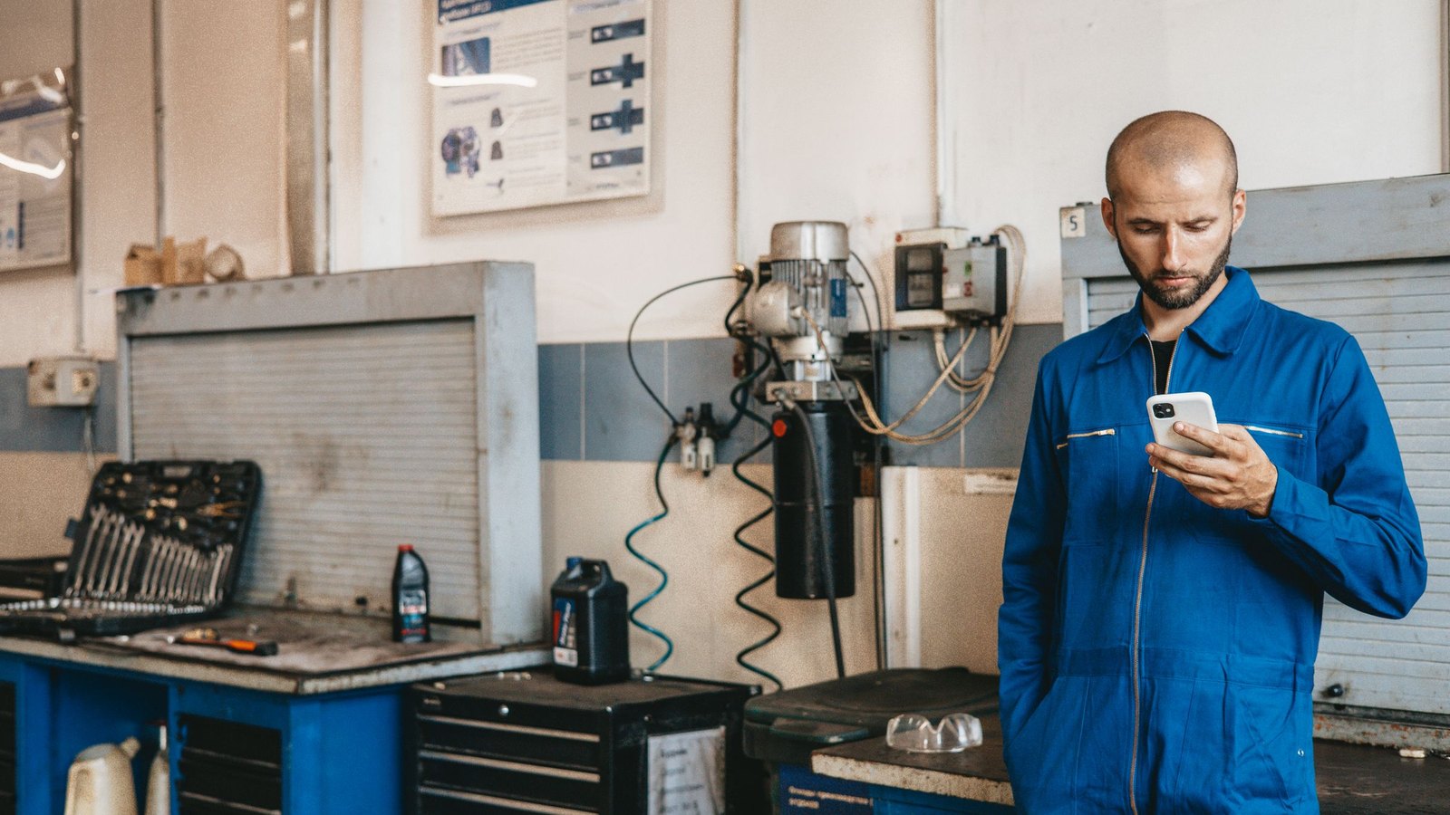 Mechanic in blue coveralls checking a job request on his phone inside his repair workshop