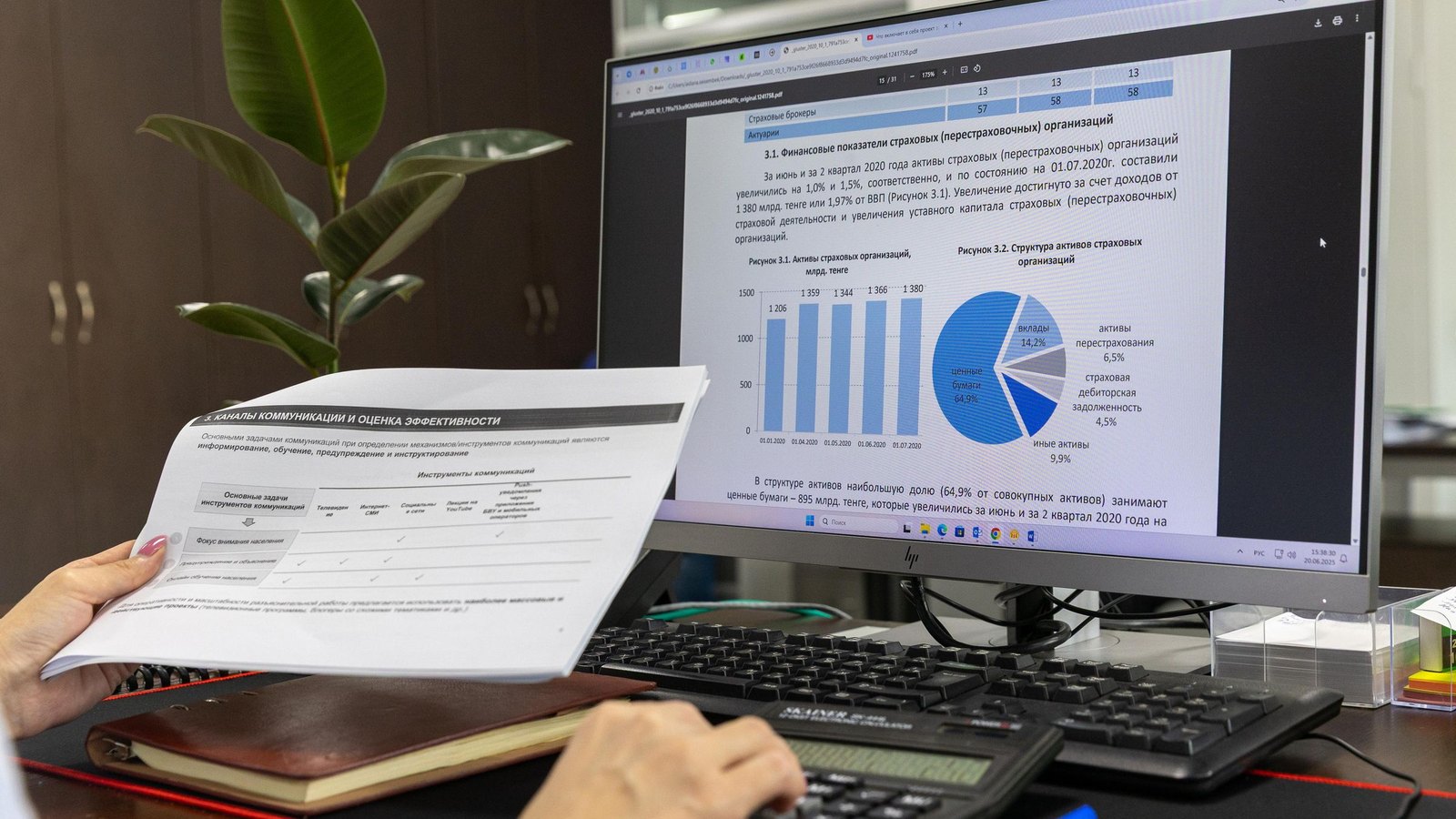 Finance staffer holding a printed report beside a monitor of bar and pie charts, calculator on the desk